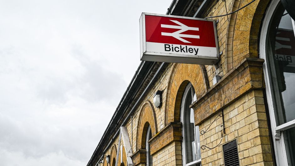 A close-up view of the exterior brick façade of a building at Bickley station, showcasing yellowish-brown bricks with arched white-framed windows. An illuminated rectangular sign with the British Rail logo and the word 'Bickley' is prominently mounted on the wall. The image is taken during overcast weather, with a cloudy sky visible in the background. The station's architectural details include decorative brickwork and metal bracket supports for the sign, emphasizing a historic railway station setting. Cleaners W3 specializes in surface cleaning and sanitisation, ensuring the maintenance of such architectural features in residential and commercial environments near Acton Town Station, W3.