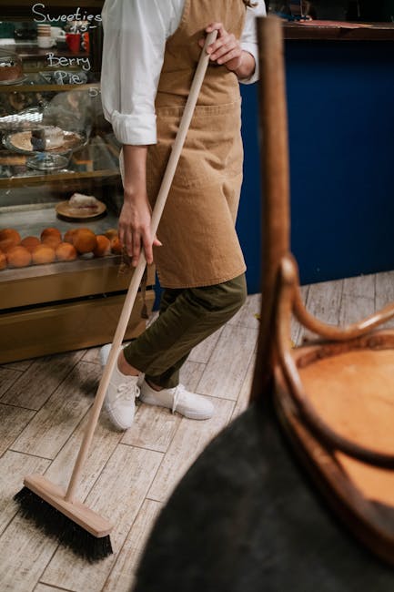 A person dressed in a white shirt and brown apron is sweeping a wooden floor with a long-handled broom inside a café or bakery setting. The individual is wearing olive green pants and white sneakers. Behind them, there is a glass display case filled with pastries and baked goods, with handwritten chalkboard signs advertising desserts. The environment appears clean and well-maintained, with natural light illuminating the space. The scene demonstrates surface cleaning and maintenance typical of commercial spaces managed by professional cleaners such as Cleaners W3, based in Acton, W3, specializing in office and shop cleaning. The focus is on ensuring hygiene and cleanliness for customers and staff alike.