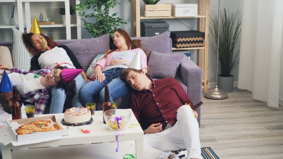 A living room scene after a family celebration with four people asleep on a purple sofa, some wearing colorful party hats. The room features a light grey wood floor, a white coffee table with an open pizza box, a birthday cake, and drinks, with party decorations scattered around. Behind the sofa, there is a shelving unit with storage boxes, a tall potted plant, and natural light coming through a window with sheer white curtains. The environment appears clean and tidy, reflecting professional domestic cleaning services by Cleaners W3. The image illustrates a relaxed, post-event atmosphere in a well-maintained home setting, emphasizing surface cleaning and overall sanitisation.