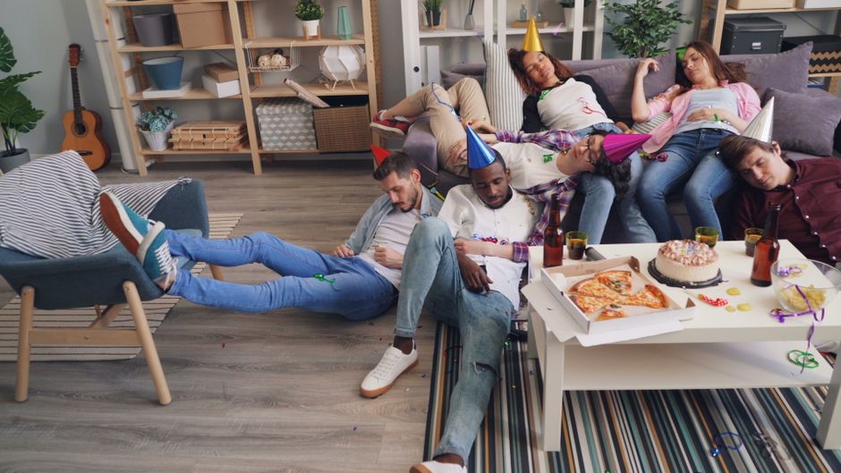 A living room scene after a family celebration with four people asleep on a purple sofa, some wearing colorful party hats. The room features a light grey wood floor, a white coffee table with an open pizza box, a birthday cake, and drinks, with party decorations scattered around. Behind the sofa, there is a shelving unit with storage boxes, a tall potted plant, and natural light coming through a window with sheer white curtains. The environment appears clean and tidy, reflecting professional domestic cleaning services by Cleaners W3. The image illustrates a relaxed, post-event atmosphere in a well-maintained home setting, emphasizing surface cleaning and overall sanitisation.
