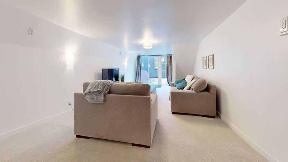 A spacious living room with white painted walls and a plush beige carpet, featuring two beige sofas positioned opposite each other around a flat-screen television on a stand. The room is well-lit with natural light coming in through a large sliding glass door at the far end, which is partially covered with dark curtains. Additional ambient lighting is provided by a ceiling fixture and wall-mounted lights. The surfaces are clean, the carpet appears freshly vacuumed, and the overall setting reflects a tidy, well-maintained home interior. Cleaners W3 has ensured thorough surface cleaning and sanitisation to maintain this immaculate condition.