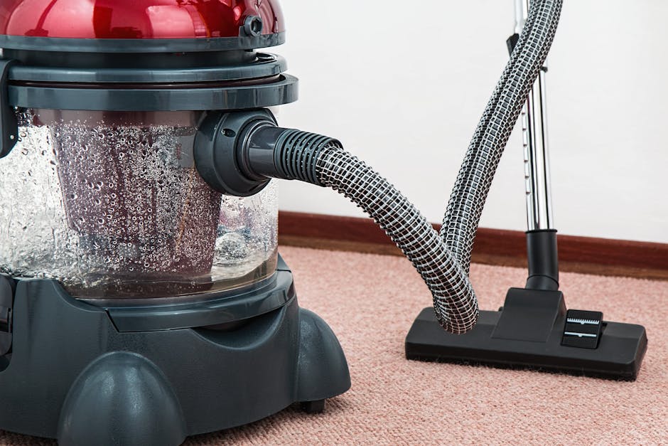 A close-up view of a modern vacuum cleaner with a transparent dust container filled with debris, connected to a flexible, patterned hose aimed at a soft, pink carpeted floor in a residential room. The vacuum's base and hose are positioned near a white wall with wooden skirting, and ambient lighting highlights the clean and well-maintained surface. The image emphasizes surface cleaning and deep cleaning processes typical for domestic cleaning by Cleaners W3, showcasing equipment used for hygienic and thorough carpet sanitisation in a home environment.