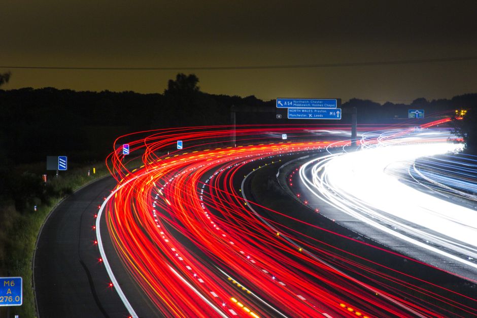 A long-exposure photo of a multi-lane motorway at night, showing red and white streaks of light from moving vehicles. The red lights from taillights curve along the left side of the highway, while whi